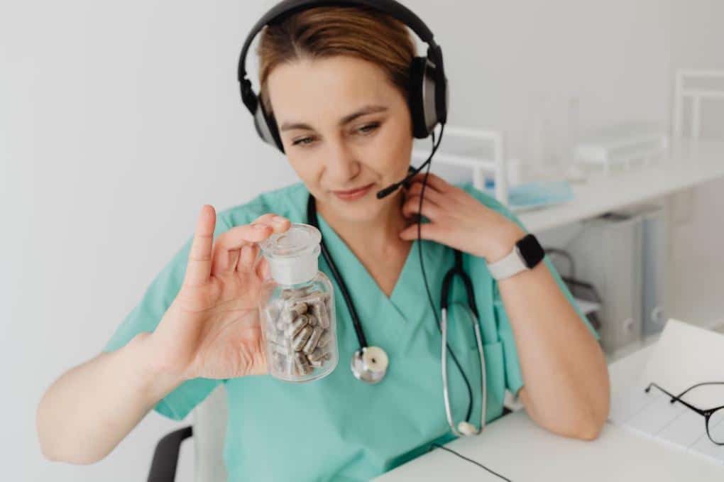 Doctor wearing headphones holding a pill bottle while speaking to a patient during a virtual consultation on a laptop.