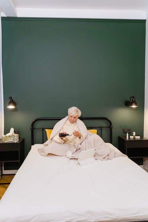 An elderly woman sitting in bed, holding a glass of water and taking her daily maintenance medication from a small pill cup.