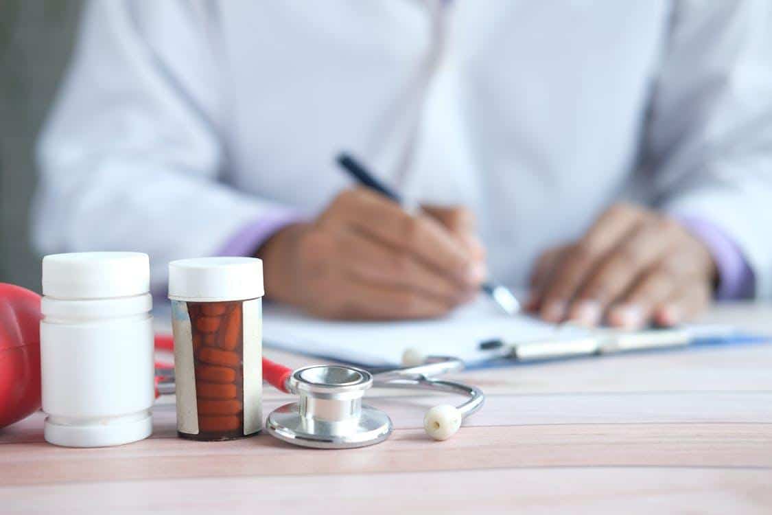 Close-up of medicines and a stethoscope on a table with a blurred doctor in the background writing a prescription.