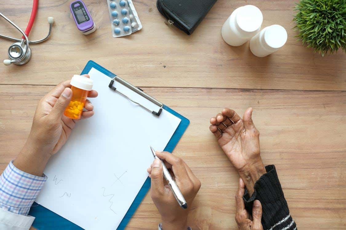 Cropped hands of a patient and a doctor, with the doctor holding a prescription pill bottle over a notepad.