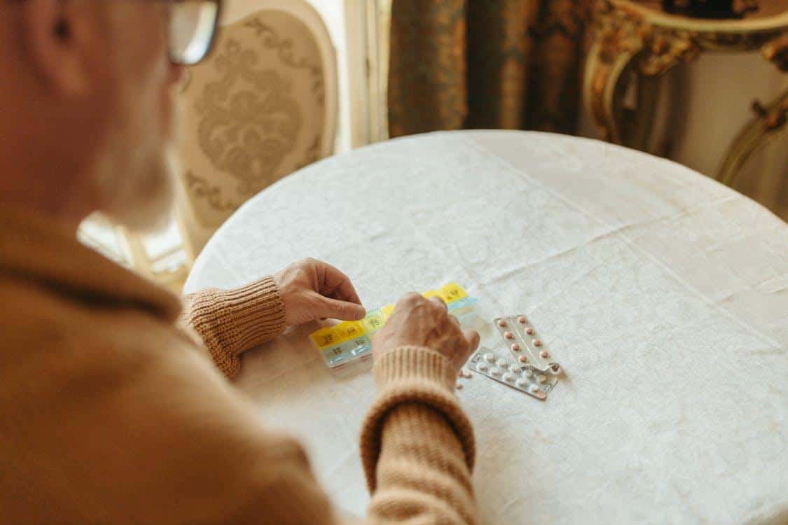 A person wearing a cozy knitted sweater sits at a table and carefully organizes various pills into a clear plastic weekly medication planner.