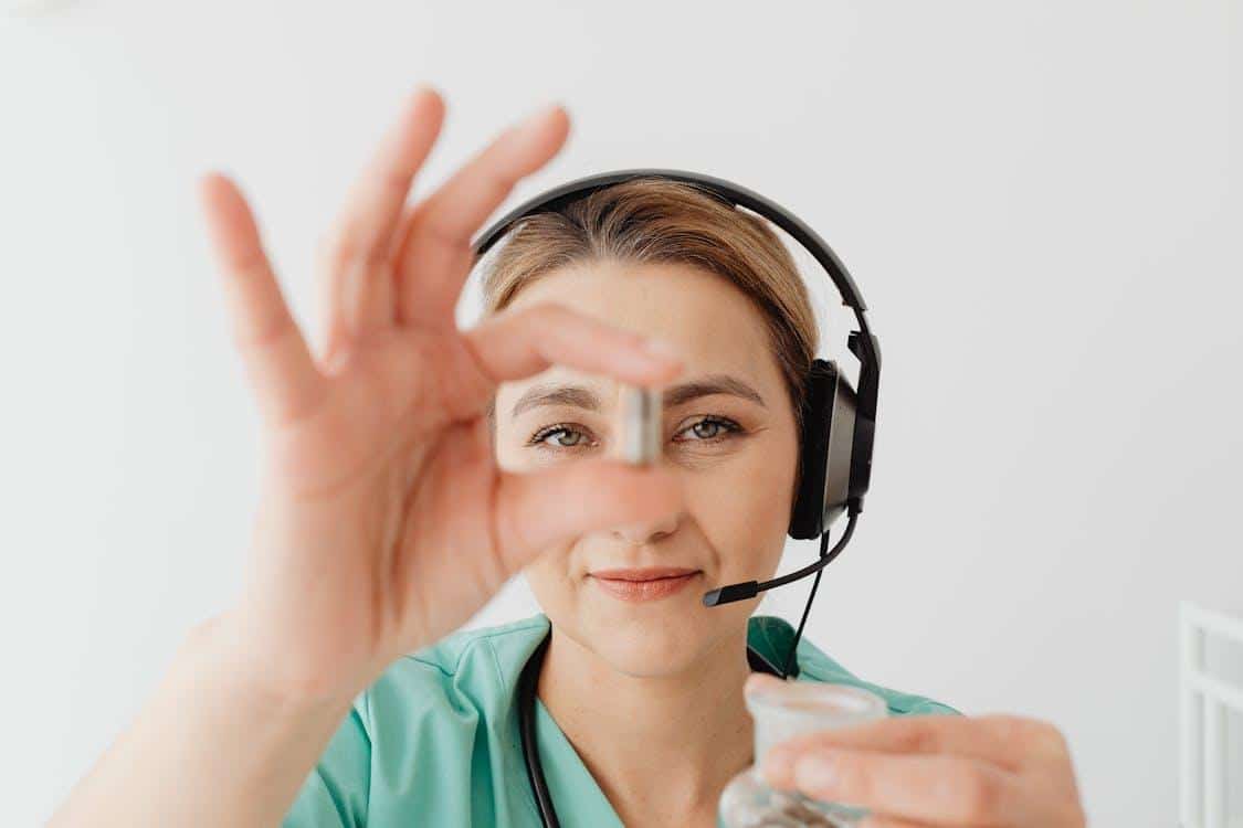 A healthcare professional wearing a headset and white coat holds a single capsule between her fingers while speaking during a virtual consultation.
