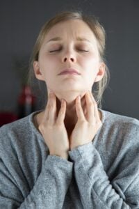 A woman with her eyes closed touches the front of her neck with one hand, expressing physical discomfort.