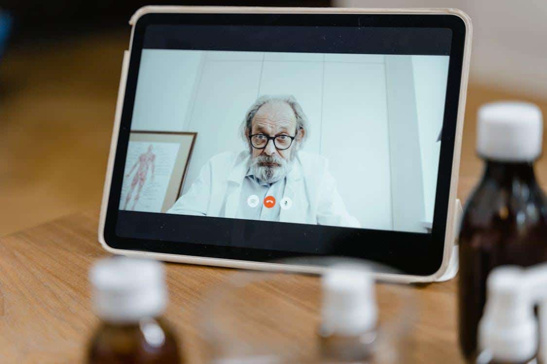 A smiling doctor wearing a white coat and stethoscope appears on a laptop screen during a secure telehealth video call.