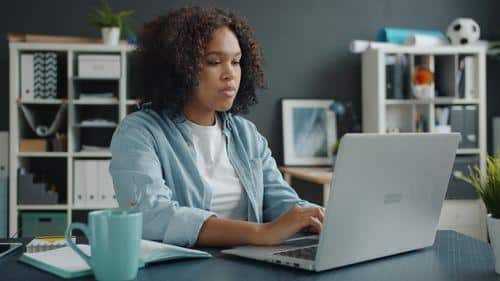 Woman sitting at her desk working remotely from home while using a laptop in a home office.